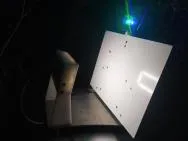 Cette photo du dispositif d’étude montre une planche blanche rectangulaire sur la droite, éclairée dans la nuit, positionnée presque verticalement sur un support portant également une caméra dirigiée vers la planche. Des petites tâches sombres sont visibles sur la planche.