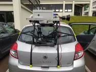Photo du dispositif d’étude consitué d'une caméra dans un boitier blanc posée sur un châssis, lui-même maintenu par de nombreuses sangles sur le capot du coffre d’une voiture. L’objectif de la caméra pointe vers le côté droit de l’image (le bas coté de la voie).