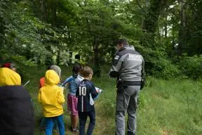 Photo d'un inspecteur de l'environnement lors d'une sensibilisation auprès d'un groupe d'enfants dans une forêt. On voit l'inspecteur de dos, habillé en tenue de terrain et armé, sur le dos de sa veste est écrit "Police Environnement" et un logo "OFB" est sur son bras gauche. 6 enfants sont à ses côtés, ils l'écoutent et ont de quoi écrire dans leur main, pour noter les indices de leur jeu d'enquête. On voit des arbres verts et de l'herbe.