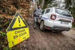 Un agent de l'OFB marche à côté de son véhicule marqué "Police de l'environnement". Au premier plan figure un panneau portant la mention "Chasse en cours"