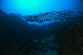 Photo d'un fond marin sombre, le sol est noir et montre  comme 2 bosses entre lesquelles un filet de pêche dérivant s'est accroché, tendu entre les 2. Il mesure plusieurs mettres de long, un plongeur passe en desous.