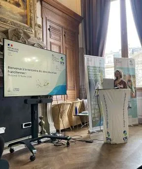 On voit un écran sur lequel est écrit bienvenue à la rencontre des aires éducatives franciliennes, sur fond blanc avec logos OFB, ministère éducation nationale, graine ile-de-france et académie du climat. une présentatrice a le micro dans les mains, elle est devant un mange-debout qui a un logo OFB et derrière elle on devine deux kakémonos représentant les aires éducatives avec des enfants et un oiseau, le chardonneret élégant. On est dans une grande salle avec du parquet en biseau, une grande porte en bois