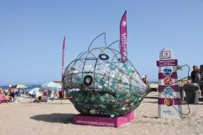Photo d'une plage fréquentée en été, avec un gros poisson en grillage rempli de bouteilles et autres déchets, accompagné d'un panneau du département de l'Hérault "Zéro plastique avec nos poissons collecteurs" et " #poissonsgloutons ".