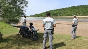 3 agents en uniforme au bord de la rivière qui regardent un adolescent sortir de l'eau. Le scooter du jeune est garé au bord de l'eau