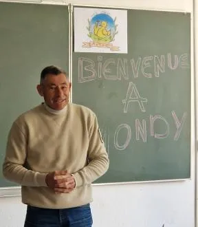 M. François SARANO intervient dans une classe scolaire. Sur un tableau à craie derrière lui, on peut lire "Bienvenue à Mondy"