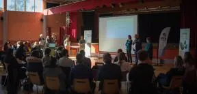 Dans une grande salle, des spectateurs écoutent une présentation sur le dispositif des Aires Terrestres Éducatives en Auvergne