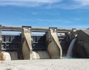 Photo d'un barrage fermé vu de l'aval vers la barrière, là où elle peut s'ouvrir : des piles de béton tenant des pans amovibles/portes, actuellement fermées, avec un petit exutoire d'eau sur la droite. Au premier plan, un peu d'eau au pied du barrage et une surface en graviers.