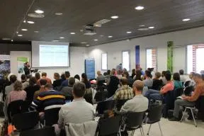 Photo d'un séminaire en salle : une projection est faite sur écran en fond d'image, et une centaine de personnes assises, de dos, regardent l'écran et une personne dedout un micro à la main.