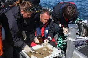 Photo : 2 hommes et une femme sur un bateau sont penchés au dessus d'un bac en plastique blanc rectangulaire contenu des sédiments (type sable beige). 