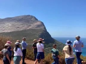 Photo d'un groupe de 10 personnes dos à l'objectif, face à un point de vue élevé, regardant au loin vers la droite le paysage. En arrière plan une montagne est visible et le ciel bleu.